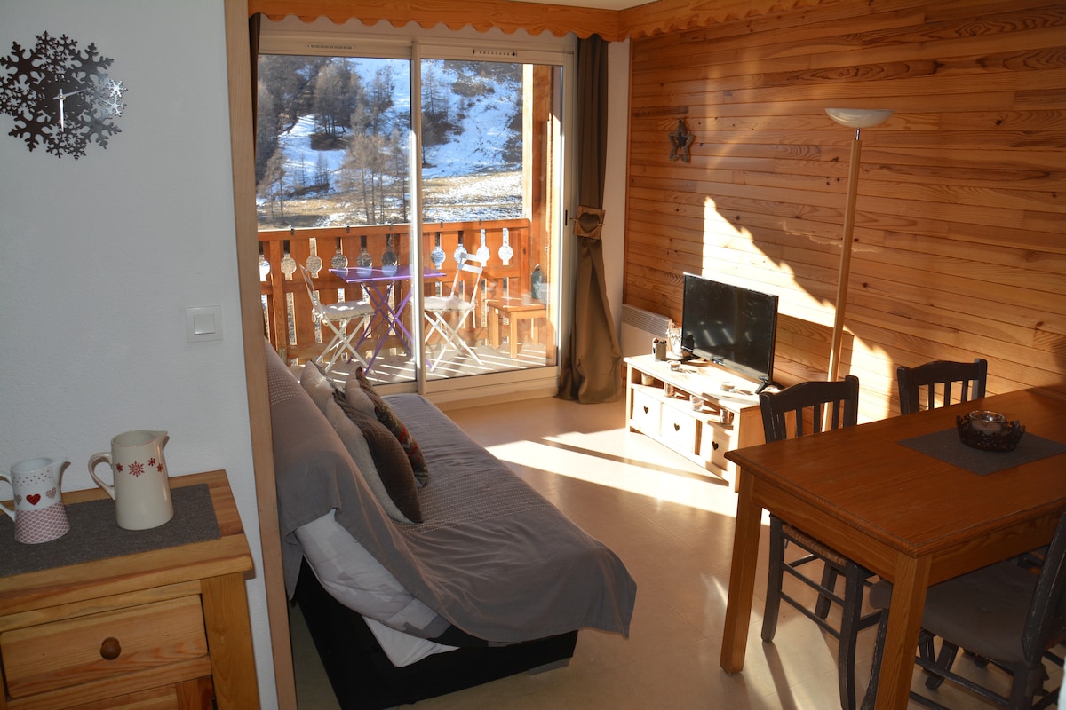 A living area featuring warm wood paneling and natural light. A spacious sofa bed faces a small wooden table and chairs. A television is situated on a console. A balcony with outdoor furniture offers views of the snowy landscape beyond.