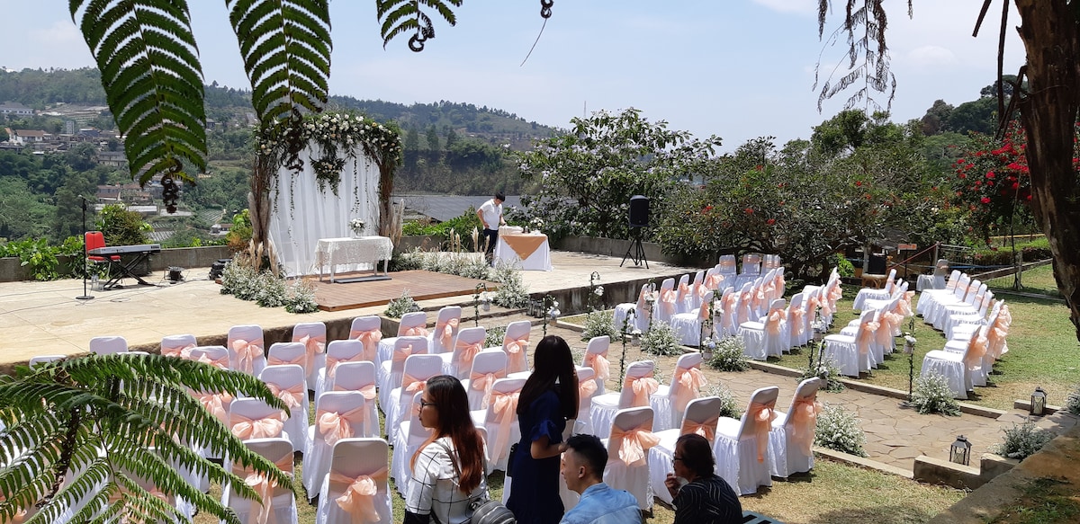 An outdoor wedding venue is set up with rows of white chairs adorned with peach sashes, facing a decorated stage. Lush greenery and floral arrangements complement the scenic backdrop of a hill, while a decorated table is positioned at the front for the ceremony.