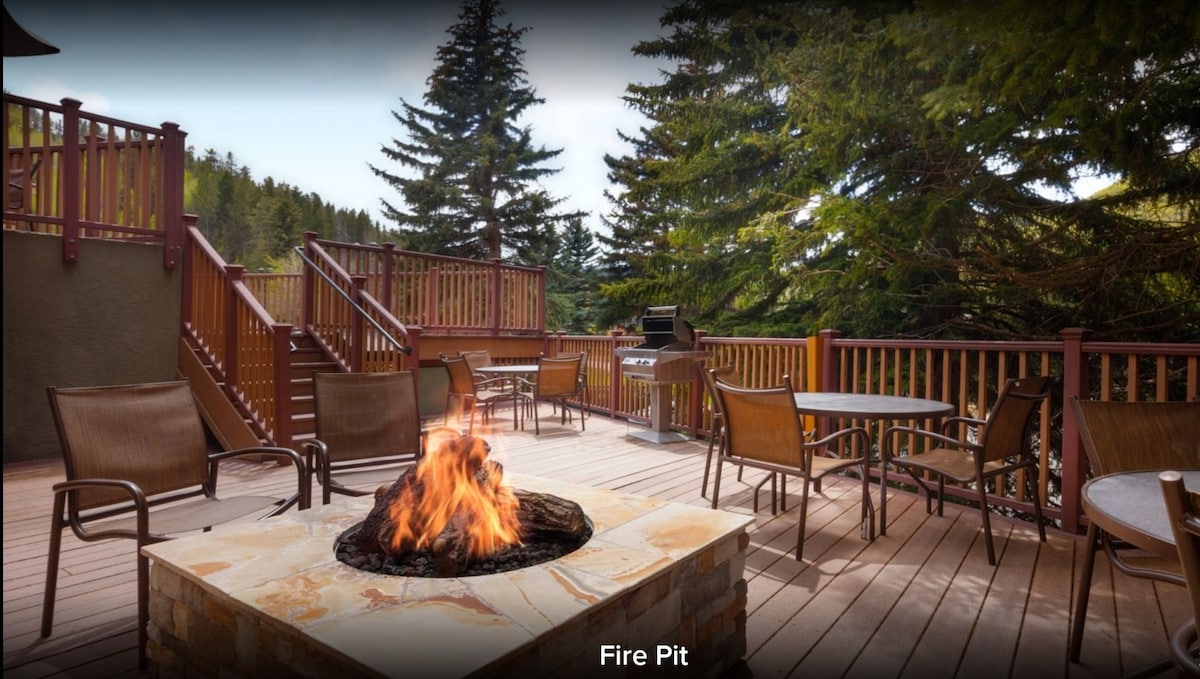 An outdoor area features a stone fire pit, surrounded by several metal chairs and tables. Wooden railings and steps lead from the elevated deck to the surrounding landscape of trees, creating a cozy space for gatherings.