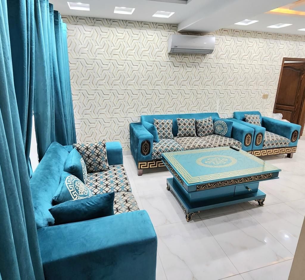 A stylish living area is furnished with multiple plush sofas in shades of blue, accented by patterned cushions. A decorative coffee table occupies the center. Bright curtains frame the windows, and the walls feature a geometric design that complements the overall decor.