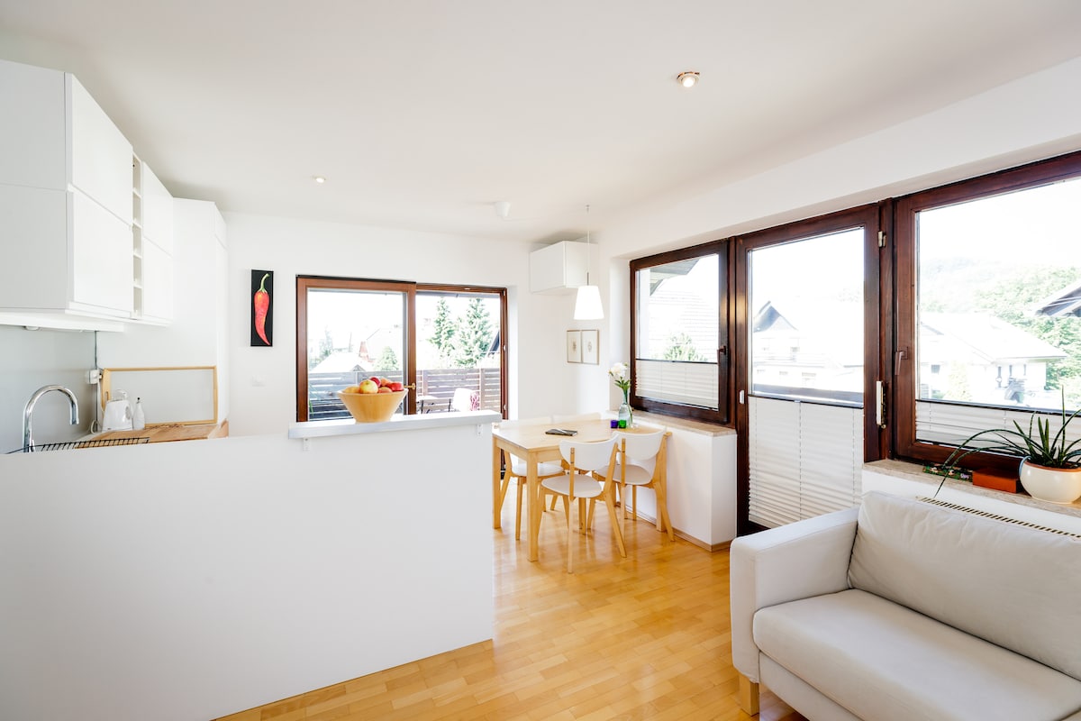 An open-plan living area features large windows inviting natural light and offering views of the surrounding landscape. A white kitchen is visible, complemented by a dining table with seating for four. A comfortable sofa is positioned nearby, and plants add a touch of greenery.