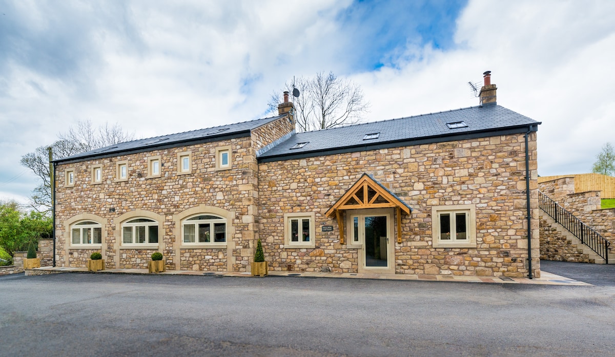 The exterior of a stone-built cottage is displayed, featuring a slate roof and large arched windows. Decorative planters flank the entrance, and a wooden overhang provides shelter. The property is set within a spacious area with gray pavement leading to an upper section, partially visible.