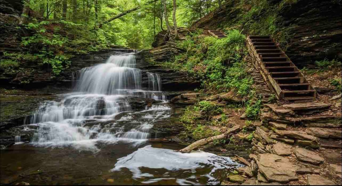 A serene natural scene features a cascading waterfall flowing over layered rocks, surrounded by lush greenery. Wooden steps lead up the slope on the right side, inviting exploration of the tranquil setting. The calm water reflects the vibrant colors of the forest.