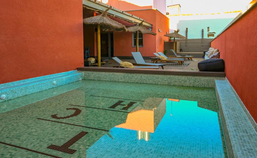 A tranquil outdoor pool area is highlighted by mosaic tiles reflecting sunlight. Comfortable loungers are positioned along the poolside, and shaded seating is provided by thatched umbrellas, creating an inviting space for relaxation.
