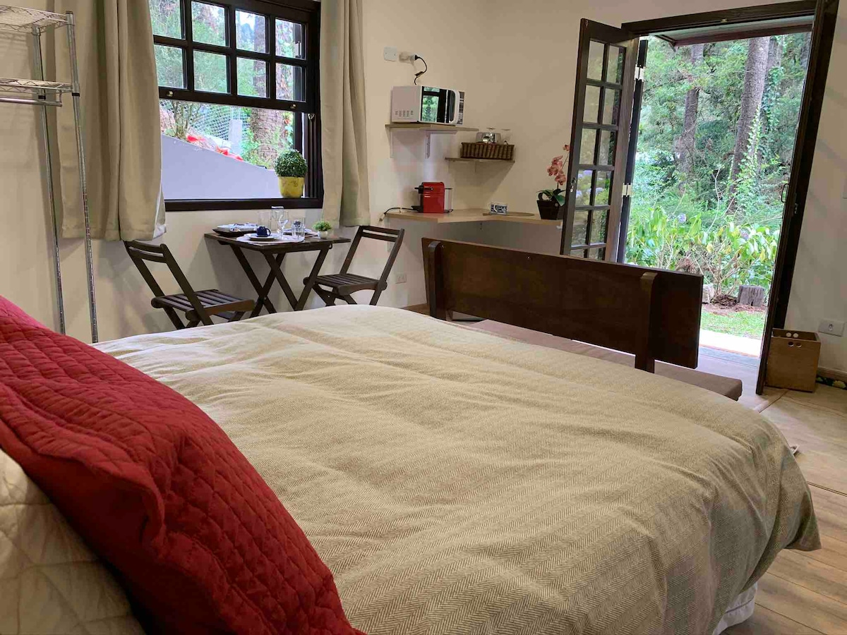 The cozy bedroom area includes a spacious bed with a light-colored blanket and a red throw. A small dining table and chairs are positioned near the window, offering views of the garden outside. A microwave and kitchenette can be seen on a shelf nearby.
