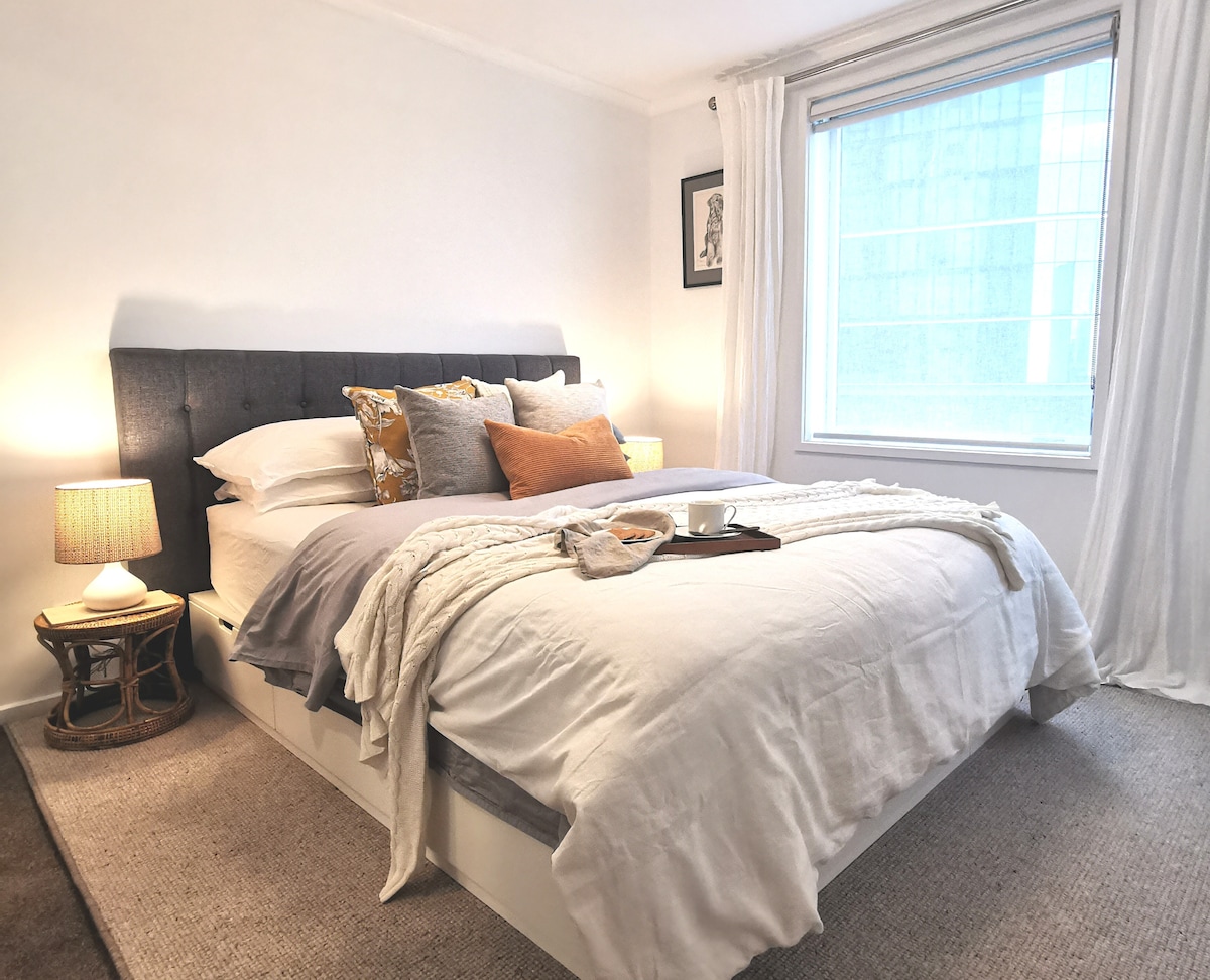 The bedroom features a spacious bed adorned with a light-colored duvet and decorative pillows. Side tables with lamps provide soft lighting, and the large window allows natural light to fill the room, revealing views of the cityscape outside.