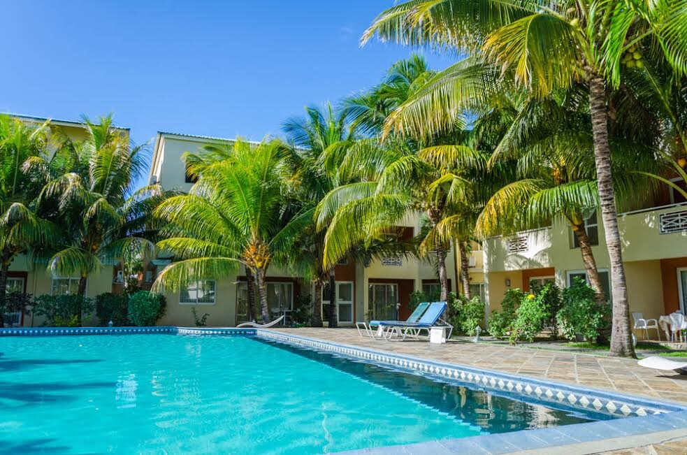 A refreshing swimming pool is surrounded by tall palm trees, providing a tropical atmosphere. Lounge chairs are positioned nearby, inviting relaxation. The two-story apartment building can be seen in the background, featuring balconies that enhance the serene environment.