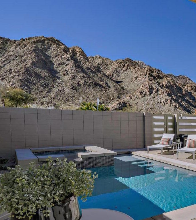 An inviting pool area is framed by a private garden wall, featuring a sleek pebble pool accompanied by a spa. Lounge chairs are positioned nearby, with the stunning backdrop of rocky mountains under a clear blue sky.
