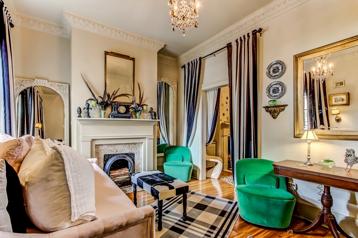 A living area showcases a beige sofa and a black and white patterned rug, complemented by two green accent chairs. A decorative fireplace is framed by ornate mirrors and a gold mirror. Stylish curtains and antique decor enhance the room's historic charm.
