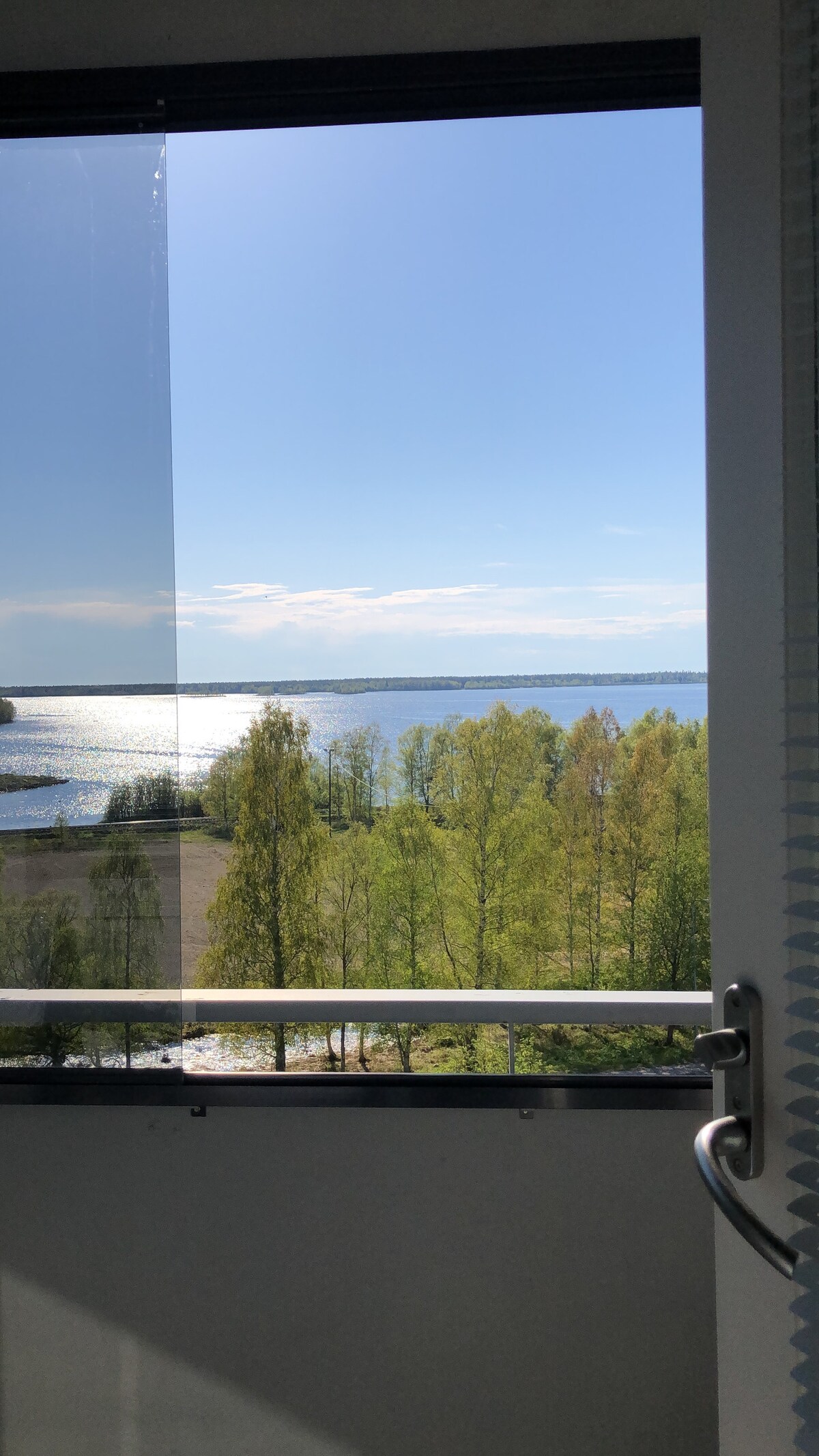 A clear view of a serene lake is visible through a glass door. Sunlight reflects off the water, surrounded by lush green trees under a bright blue sky. The scene exudes tranquility from this upper-floor perspective.