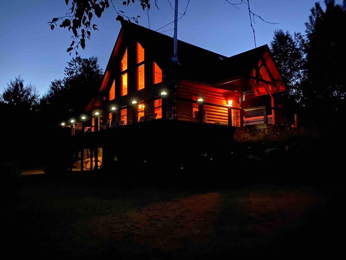The log cabin is illuminated at dusk, highlighting its rustic architecture. Large windows and a spacious deck offer an inviting appearance. Soft lighting accentuates the structure's features, showcasing the combination of modern comfort and natural elegance in the outdoor surroundings.