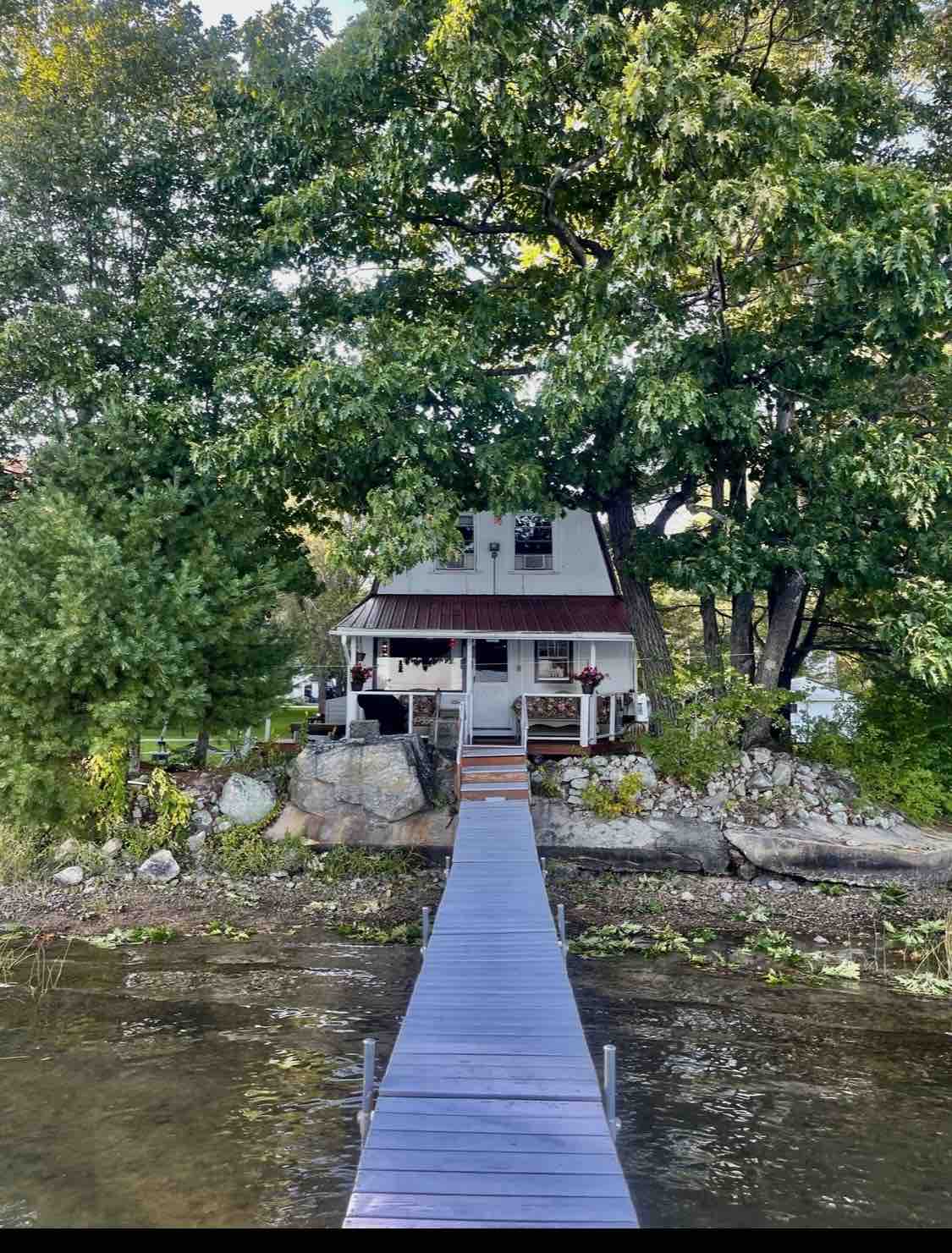 A charming cottage is nestled among trees, with a welcoming porch and a rustic roof. A wooden dock leads to the water, framed by stones and greenery, creating a serene outdoor setting. The peaceful surroundings invite relaxation and connection with nature.