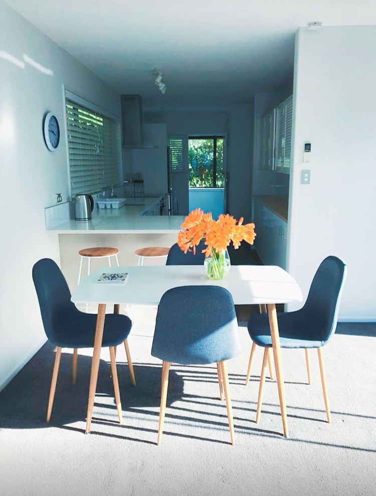 A dining area is presented with a white table surrounded by four chairs, featuring a bouquet of orange flowers as a centerpiece. Light streams in from a nearby window, revealing a bright kitchen space in the background with modern appliances.