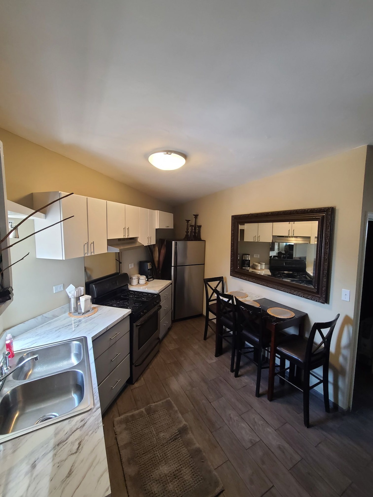 A modern kitchen is shown with white cabinetry and marble countertops. Stainless steel appliances, including a refrigerator and stove, are present. A dining area with a small round table and two chairs is positioned against the wall, complemented by a large mirror that enhances the sense of space.