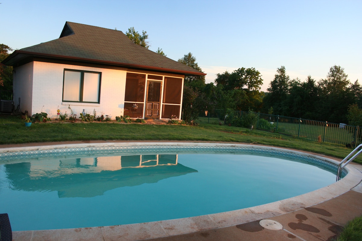 A seasonal in-ground pool is surrounded by a concrete deck, reflecting the nearby guest house and verdant landscaping. The fenced yard features a lawn with various plants, and the screen porch is visible on the house, providing a serene outdoor space.