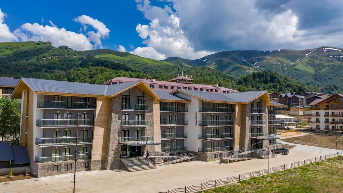 A modern multi-story building is depicted amid green mountain slopes. Constructed with a combination of stone and wood, the structure features balconies on each floor. An outdoor area is shown with gravel and pathways leading towards the entrance, creating a welcoming approach.