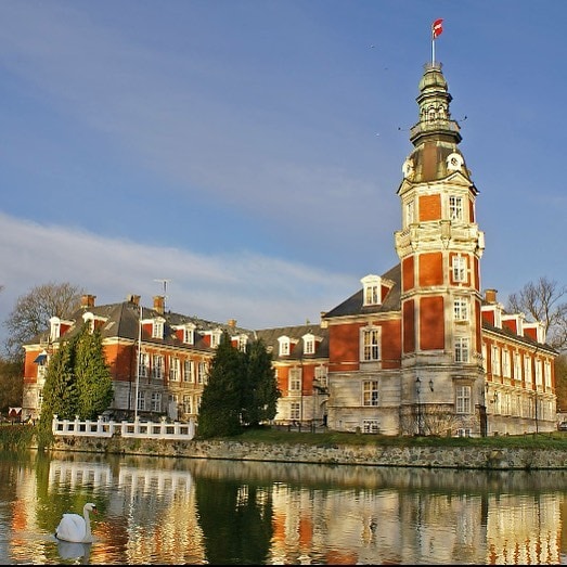A historic castle is reflected in a nearby pond, showcasing its striking red brick facade and tall tower. Lush trees surround the property, and a swan glides gracefully across the water, contributing to the serene atmosphere of the landscape.