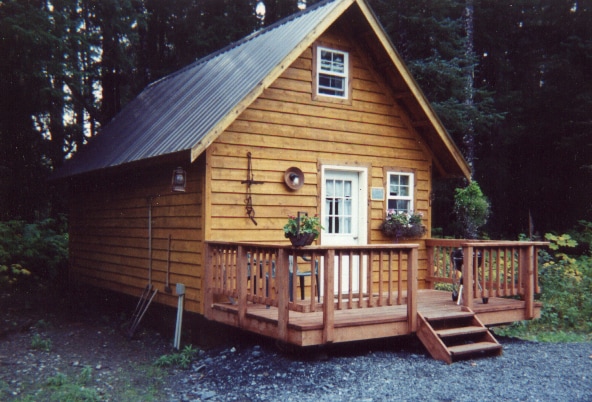 A charming wooden cabin with a gabled roof stands surrounded by trees. A front porch features a railing and steps leading to the entrance. Flower pots are placed on the railing, adding a touch of greenery to the rustic facade. Sunlight filters through the forest.