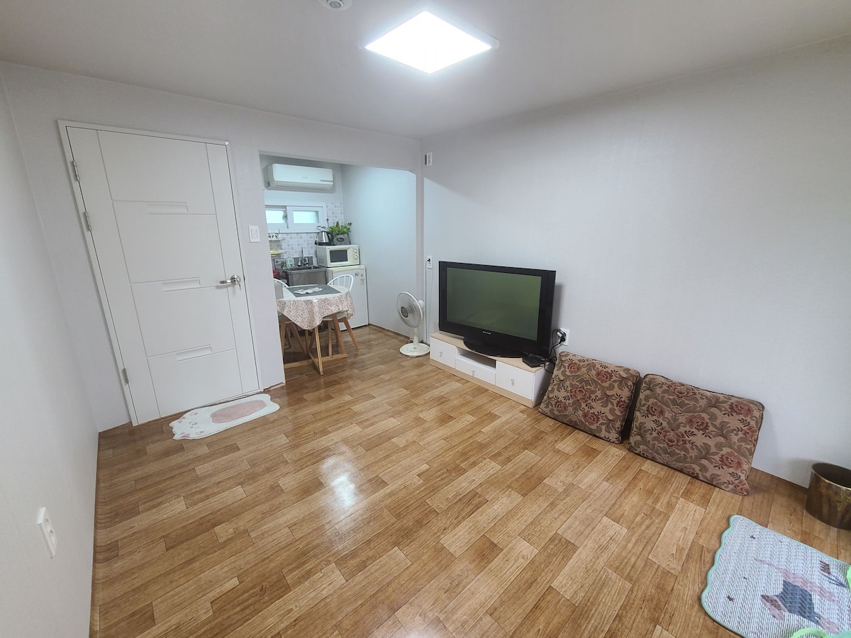 A spacious living area features a flat-screen TV positioned against a neutral wall. Two floor cushions are placed on the wooden floor, which reflects natural light from a ceiling window. A small dining area with a table and chairs is visible in the background.