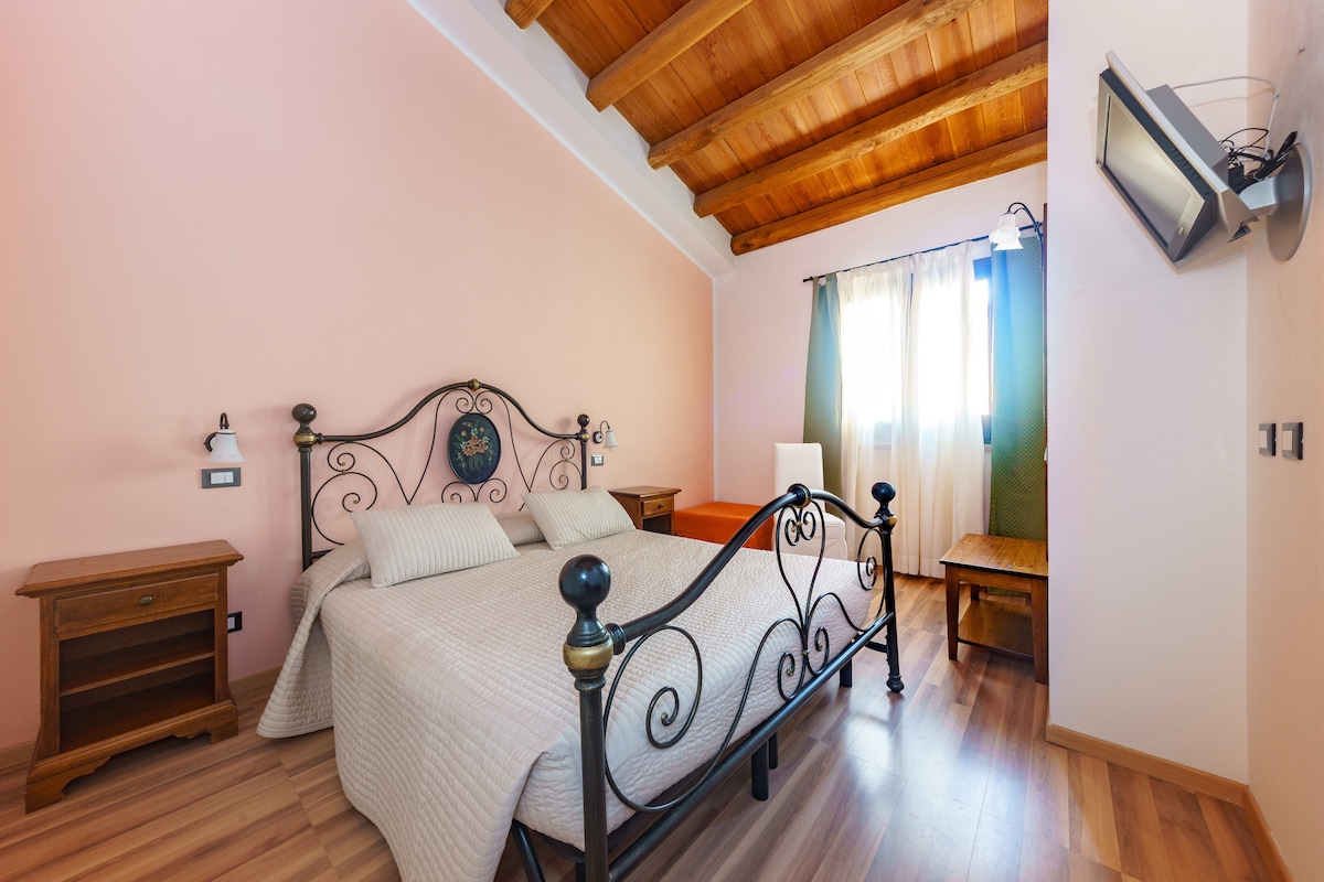 A cozy bedroom features a spacious, wrought-iron bed with light-colored bedding. Natural light enters through curtains, illuminating the room's wooden ceiling and warm peach walls. Nightstands are placed on either side of the bed, while a wooden table is positioned near a window.