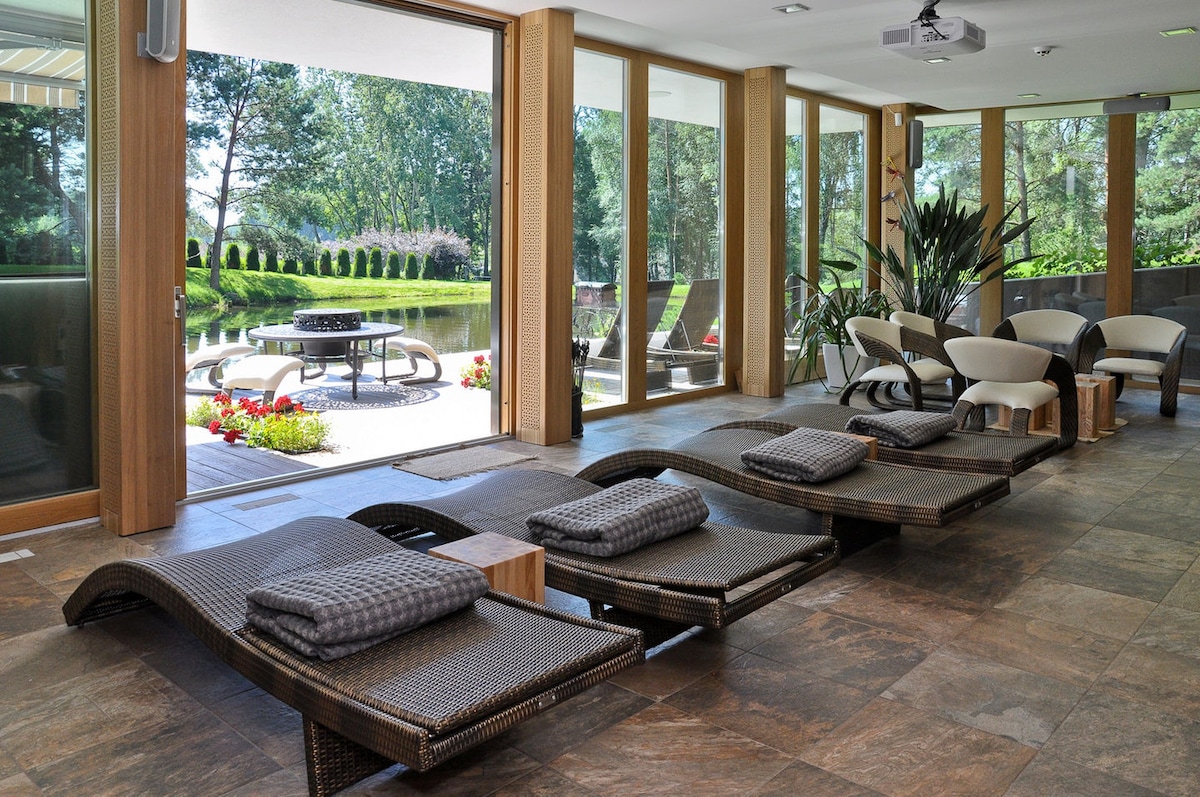 A calming indoor relaxation area features elegant lounge chairs, each paired with a soft cushion. Natural light floods through large glass doors, providing views of the serene outdoor space and garden. The room is adorned with indoor plants, enhancing the tranquil atmosphere.
