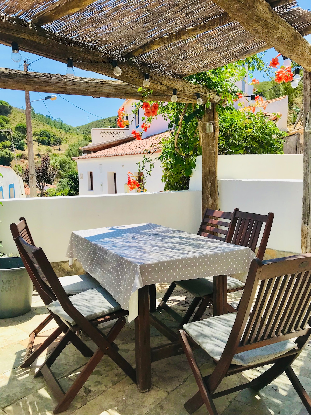An outdoor dining area is shaded by a wooden pergola adorned with vibrant flowers. A polka-dotted tablecloth covers a wooden table, surrounded by four wooden chairs. The backdrop features whitewashed walls and a glimpse of greenery from the surrounding area.