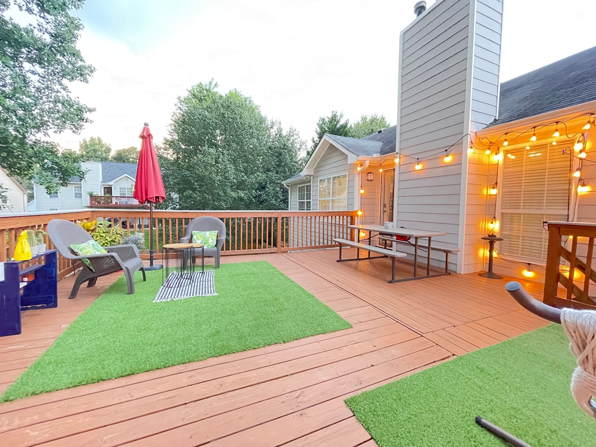 A spacious back deck adorned with string lights creates a relaxed outdoor space. Comfortable seating is provided along with a picnic table. A vibrant red umbrella adds a splash of color. The deck features areas of artificial grass for a cozy feel and easy maintenance.
