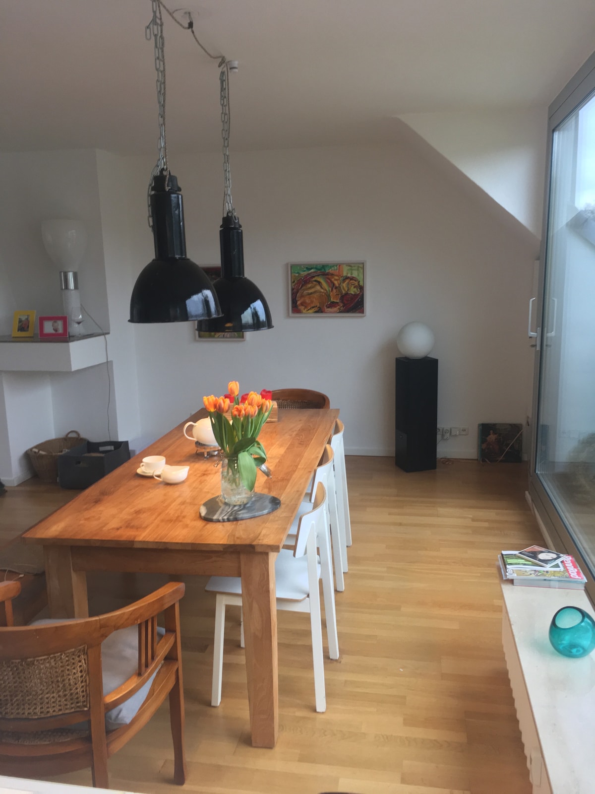 A bright dining area features a long wooden table surrounded by white chairs and two wooden armchairs. A vase of orange tulips is placed at the center. Two large black pendant lights hang above the table, while a colorful piece of art is displayed on the wall.