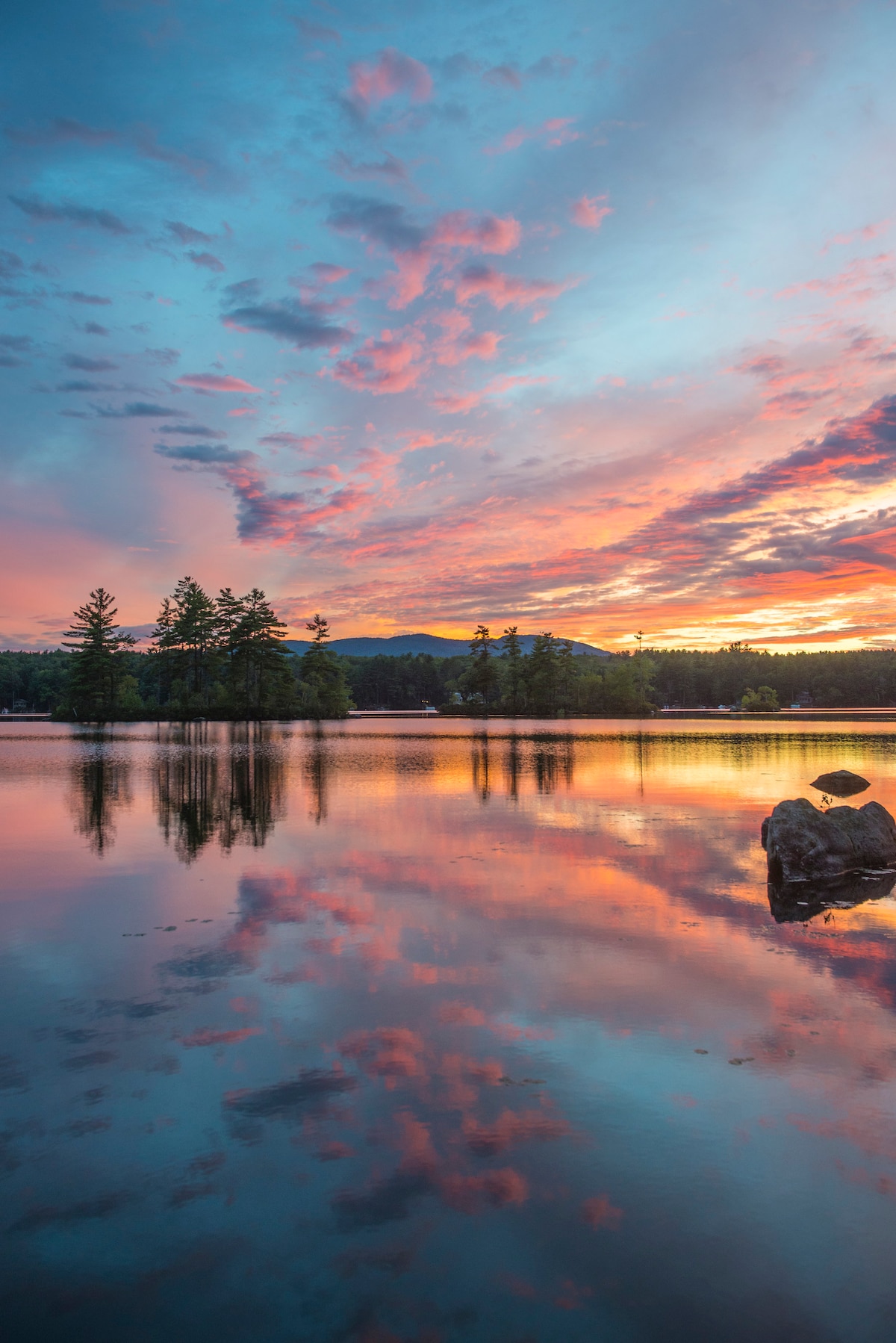 A serene lakeside scene captures the vibrant colors of a sunset reflected on calm water. Hues of pink, orange, and blue paint the sky, while silhouetted trees line the shore. A large rock in the foreground adds a natural touch to the tranquil landscape.