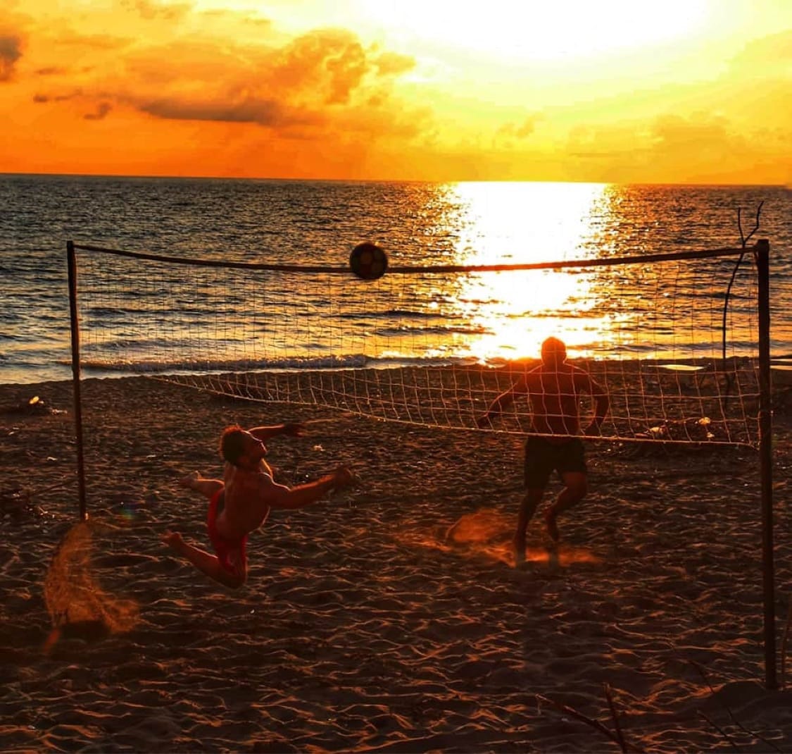 A sunset scene captures two individuals playing beach volleyball near the water. The horizon is illuminated with warm hues of orange and gold, reflecting on the calm sea. The sandy beach surrounds the net, complementing the serene atmosphere.