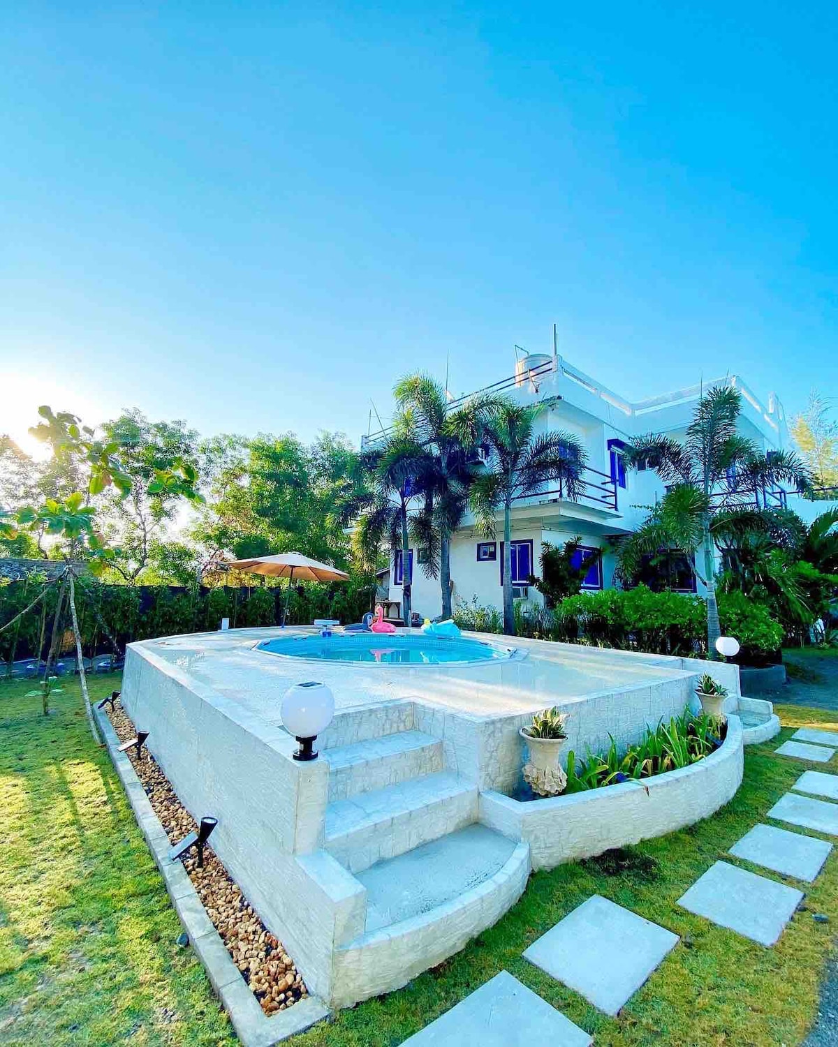 A serene outdoor area features a light-colored building surrounded by lush greenery. A small pool is situated in front, complemented by steps leading up to it. Clear blue skies enhance the tranquil setting, while varied plants and pathway stones add to the inviting landscape.