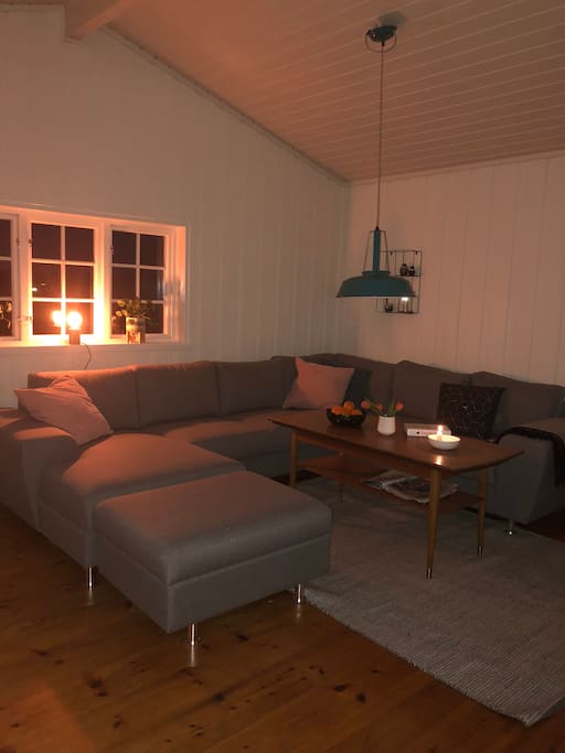 Stua med mange sitteplasser, peisovn og tv. Utgang til veranda og solnedgang i vest.