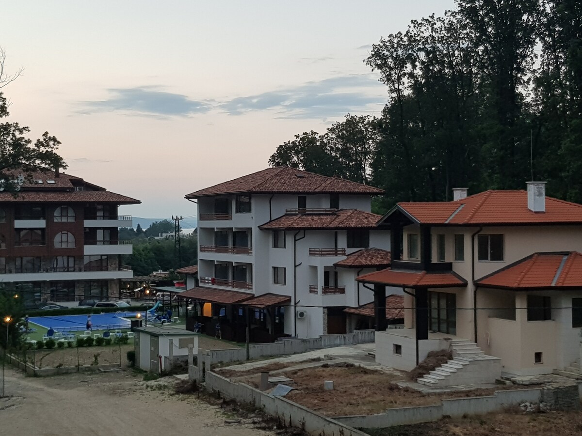 The image depicts a serene landscape featuring several buildings within an eco-friendly complex. A swimming pool is visible in front of a three-story building, surrounded by tall trees. A spacious terrace is noted on the right, enhancing the overall peaceful ambiance.