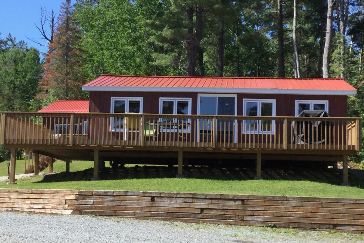 A lodge-style cottage is positioned on a raised deck with wooden railings. The structure features a vivid red roof and multiple large windows, allowing natural light to illuminate the interior. Surrounding greenery enhances the tranquil setting.