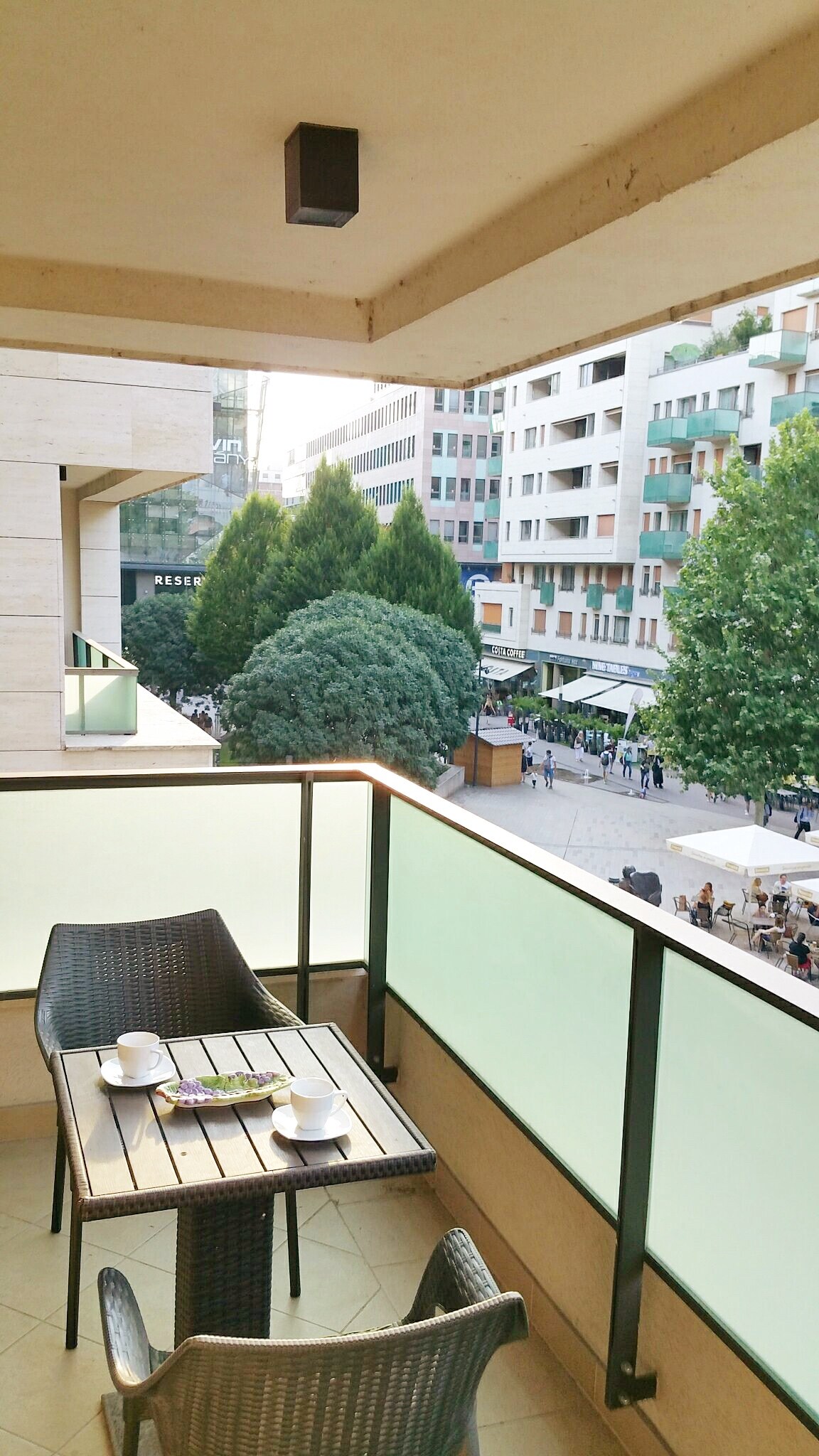 A balcony featuring a small table set with two cups and a plate is visible. The view overlooks bustling Corvin Promenade, showcasing trees and nearby restaurants, with pedestrians enjoying the vibrant atmosphere below. The glass barrier provides an unobstructed sight of the lively scene.
