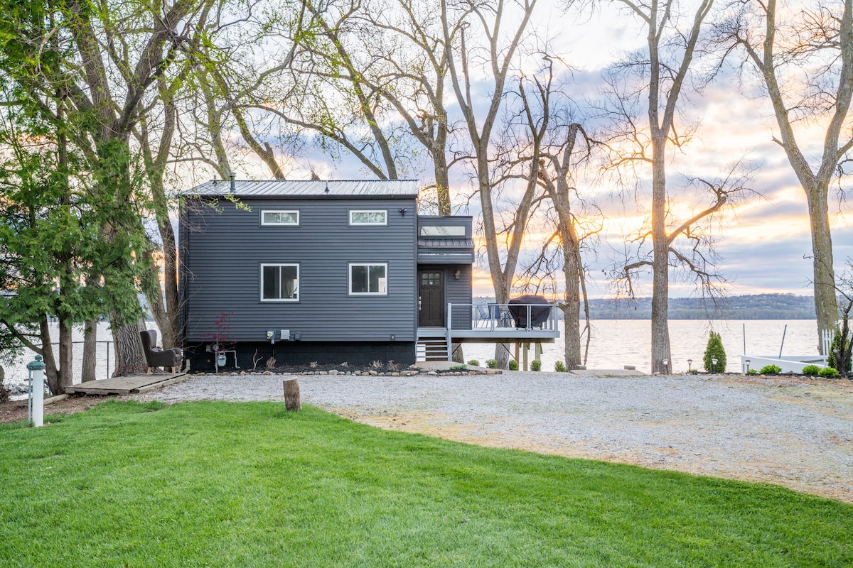 The exterior of the house is shown, featuring a modern design with large windows. Surrounding trees provide natural shade, and a well-maintained lawn leads towards the water. The sunset casts warm hues across the sky, enhancing the serene riverside setting.