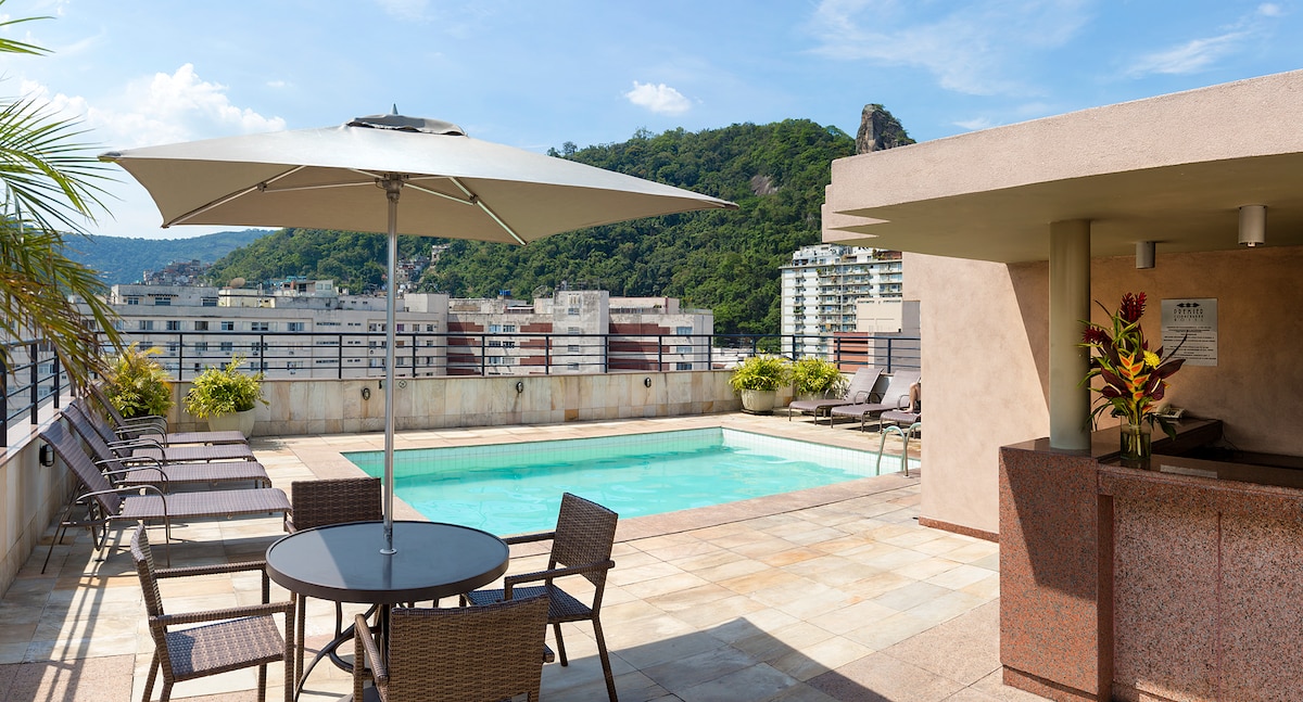 A rooftop area features a clear swimming pool surrounded by lounge chairs. A shaded table and chairs are positioned nearby. The backdrop includes lush greenery and urban structures under a bright blue sky, providing a relaxed outdoor setting.