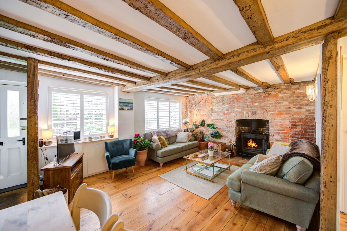 A welcoming living area is highlighted by exposed wooden beams and a rustic brick wall. Comfortable seating arrangements consist of a green couch and a light blue sofa, accented by soft cushions. A glass coffee table sits in the center, with a cozy wood-burning stove nearby, creating a relaxed atmosphere.