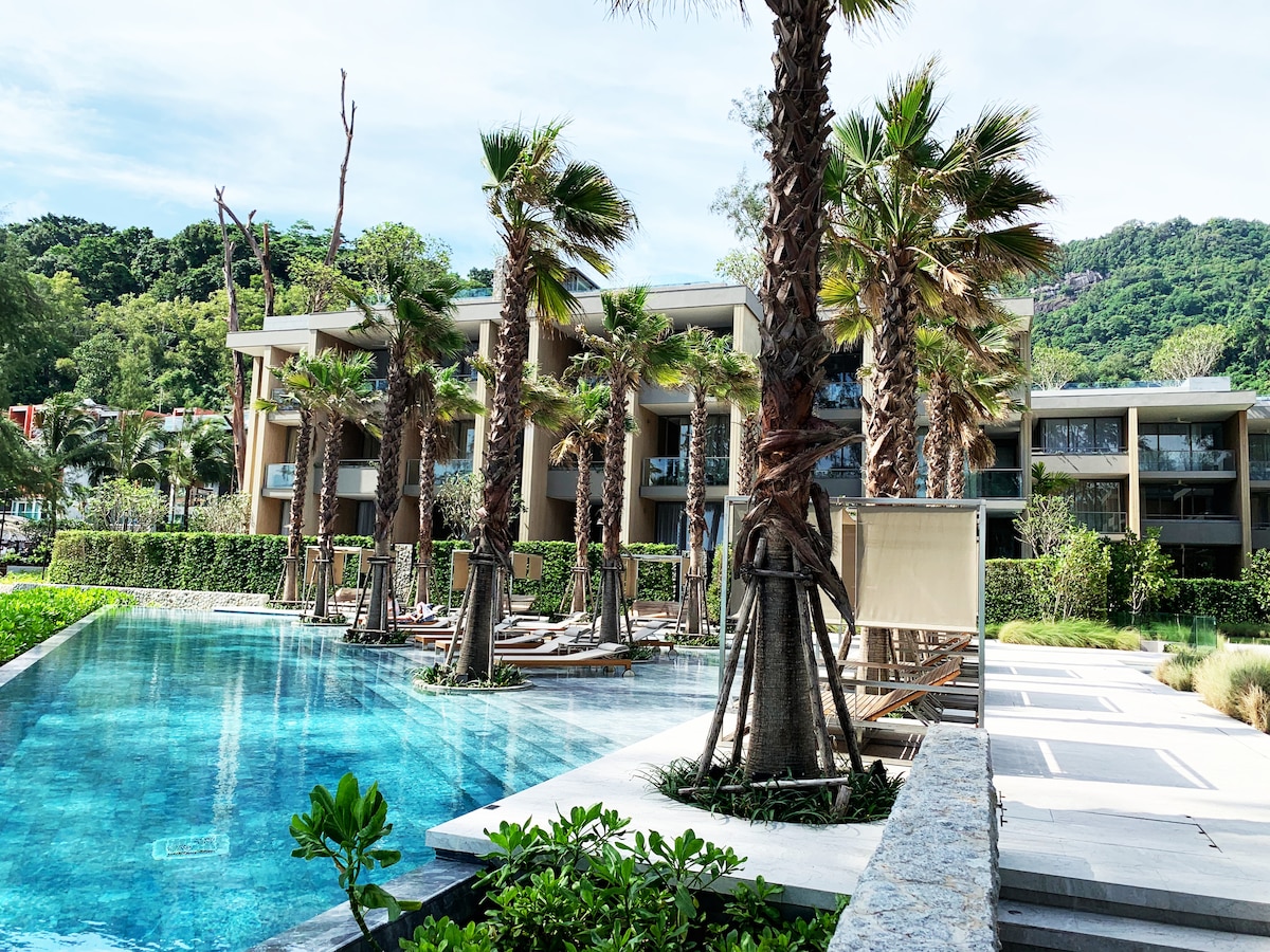 The resort features a large outdoor swimming pool, surrounded by palm trees and lush greenery. Modern buildings can be seen in the background, showcasing large windows. Sun loungers are placed around the pool, creating a relaxing atmosphere in the serene environment.