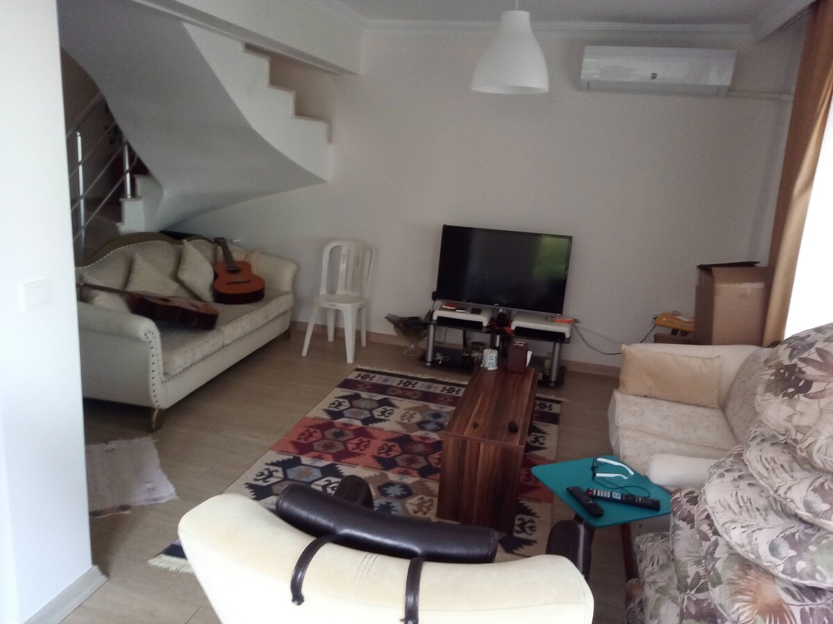 A spacious living area is depicted, featuring a light-colored sofa and a patterned area rug. A wooden coffee table is centered in the room, accompanied by a television on a stand. A guitar rests on a chair, and a staircase is visible in the background.