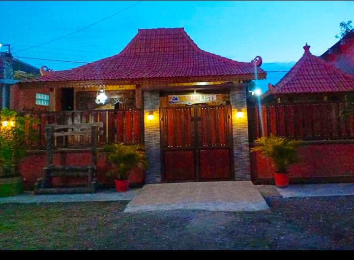 A charming entrance to a traditional-style home is highlighted by wooden doors flanked by decorative lanterns. The structure features a distinctive roof design, with potted plants positioned on either side of a stone pathway leading to the entrance, illuminated by soft evening light.