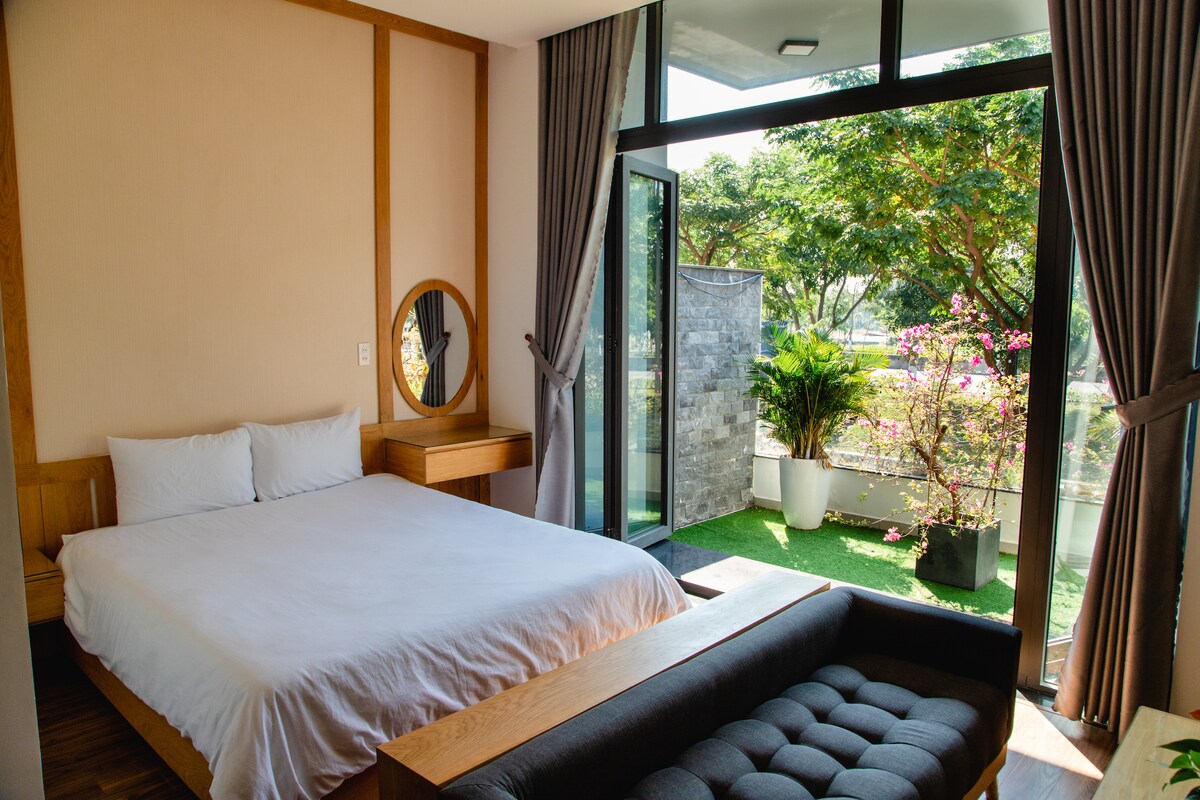 A spacious room features a queen-sized bed with a minimalistic design, complemented by a wooden desk and a round mirror. Large glass doors open to a serene outdoor area, showcasing greenery and a stone wall, allowing natural light to fill the space.