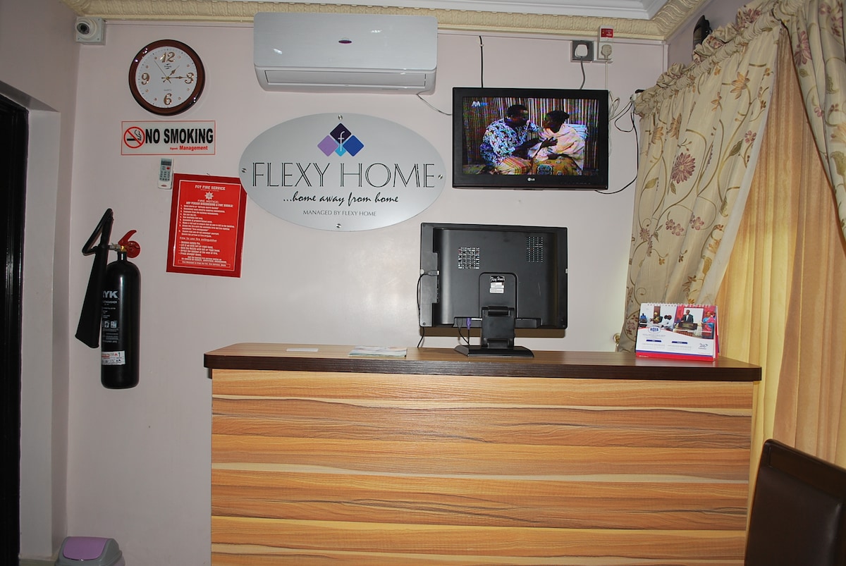 A reception area is shown with a wooden front desk and a computer setup. Above the desk, a wall-mounted flat screen TV displays content. A clock and a fire extinguisher are also visible. The space has soft lighting and decorative curtains.