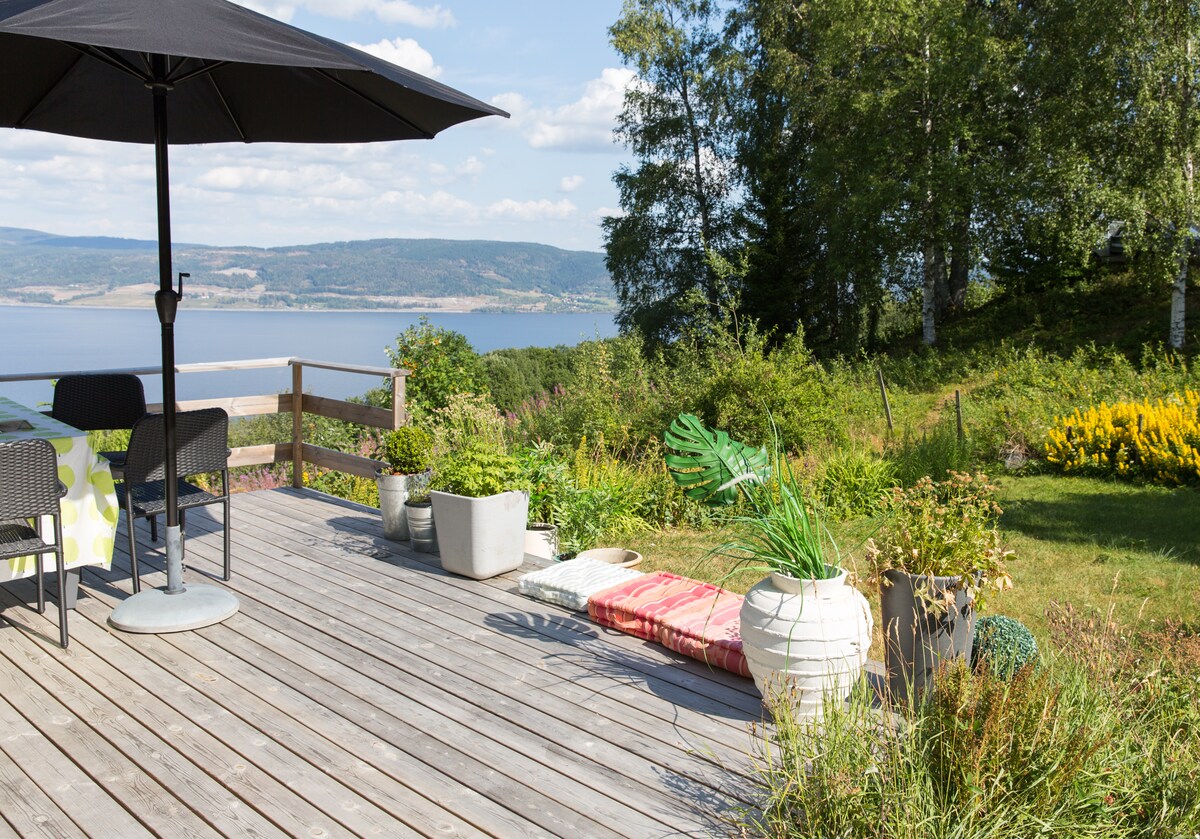 An outdoor terrace features a large umbrella providing shade over a dining table with two chairs. Lush greenery and colorful flowers frame the wooden deck, alongside potted plants and a floor mat for relaxation. The view of the lake and distant hills enhances the serene setting.
