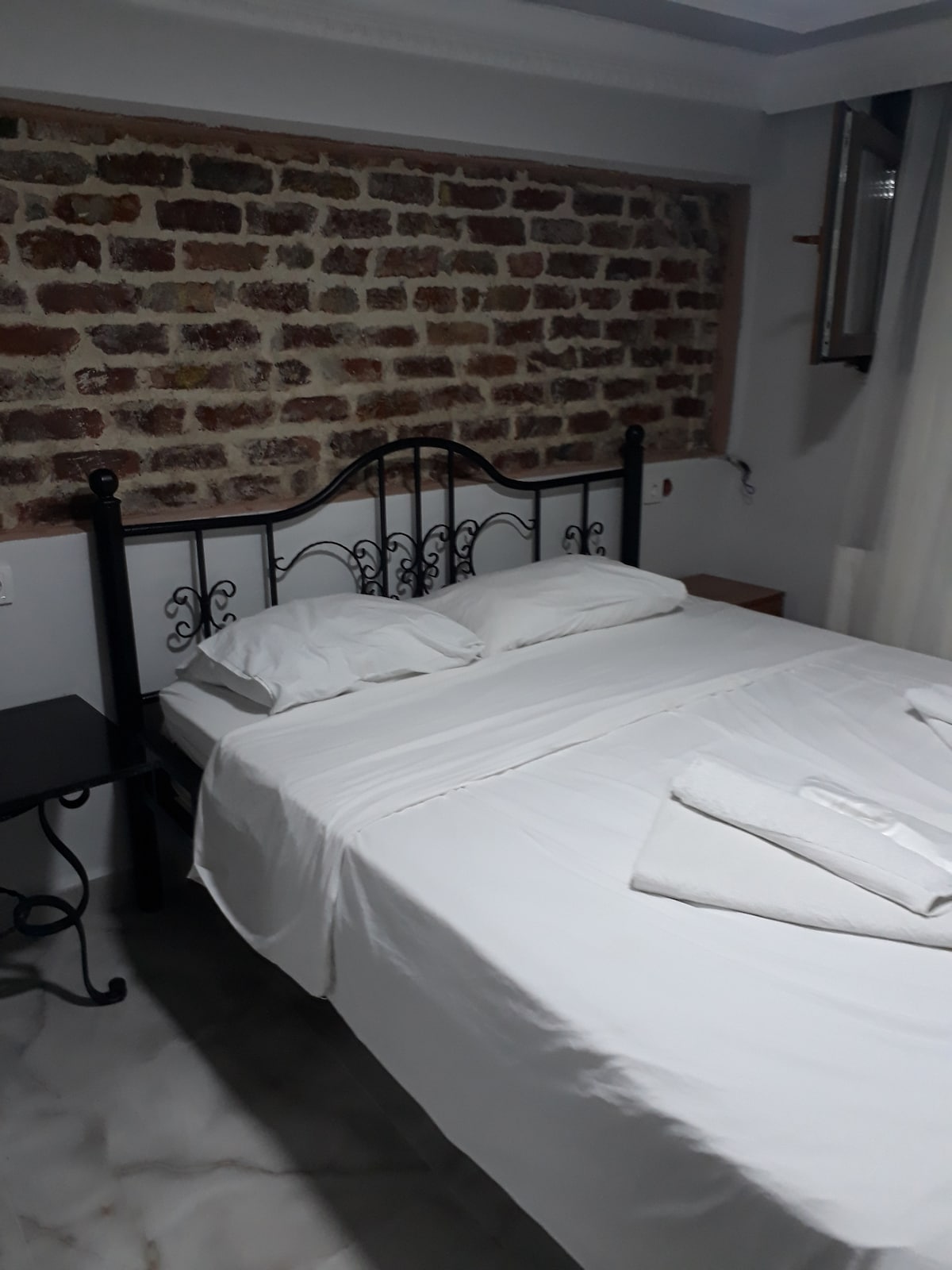 A spacious bed is positioned against a rustic brick wall, providing a warm and inviting atmosphere. Soft linens are neatly arranged, with two towels folded on the bed. A black side table is placed beside the bed, complementing the overall aesthetic.