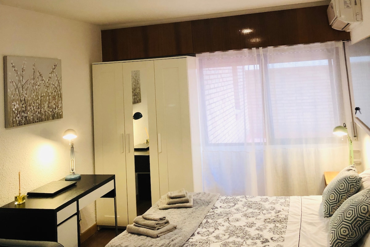 A comfortable sleeping area features a spacious bed adorned with patterned bedding. A sleek desk with a lamp is positioned near a large window, while a wardrobe provides ample storage. Soft natural light fills the room, complemented by a serene decor.