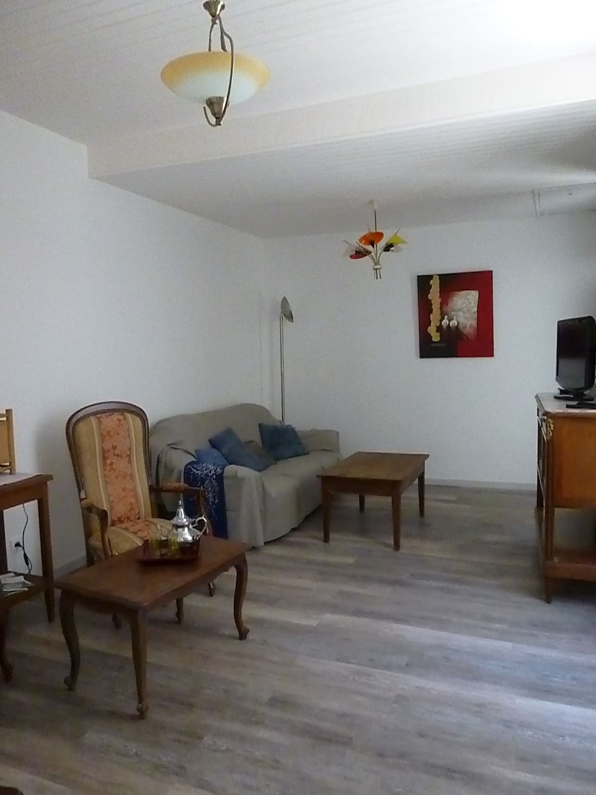 A cozy living area features a soft sofa adorned with cushions, accompanied by a wooden coffee table. An ornate chair and a vintage side table add charm to the space, while a television and a decorative artwork complete the inviting atmosphere.