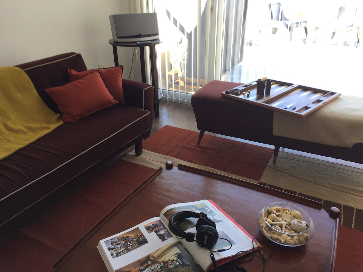 A welcoming living area is furnished with a dark sofa and a matching low table. A decorative bowl filled with snacks is positioned on the table. Nearby, a stack of magazines rests on a vibrant rug, with natural light filtering through sheer curtains.