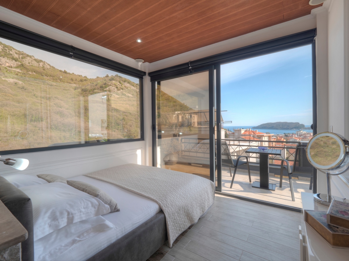 A bright bedroom features large glass doors that open to a spacious terrace, offering a view of the sea and surrounding landscape. The bed is dressed in soft linens, complemented by a neutral-colored throw. Natural light fills the room, enhancing the modern decor.