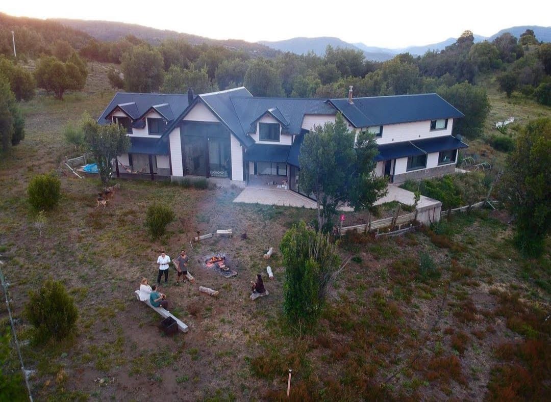 The house is positioned in a natural environment, surrounded by greenery and distant mountains. A spacious outdoor area is visible, with a fire pit and seating arrangements. The evening light enhances the peaceful setting, inviting relaxation and connection with nature.